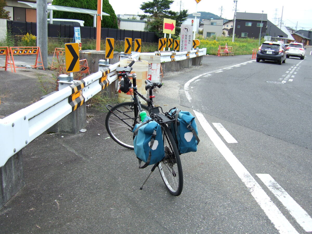 A bicycle parked in a narrow patch of asphalt between a rusted guard rail and a trafficked road.