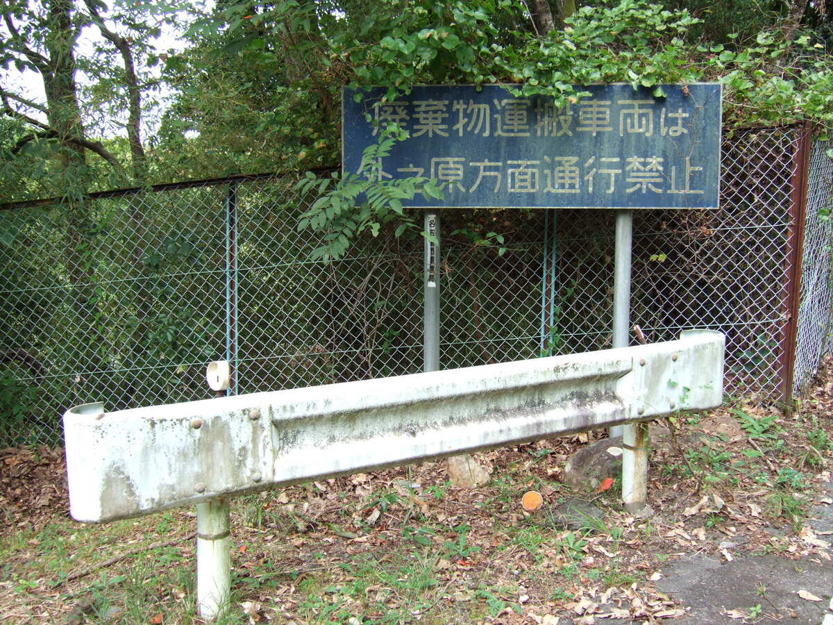 A sign standing behind a guardrail, reading (in English translation); “Trucks hauling refuse are formidden to travel through to Soto-no-Hara”