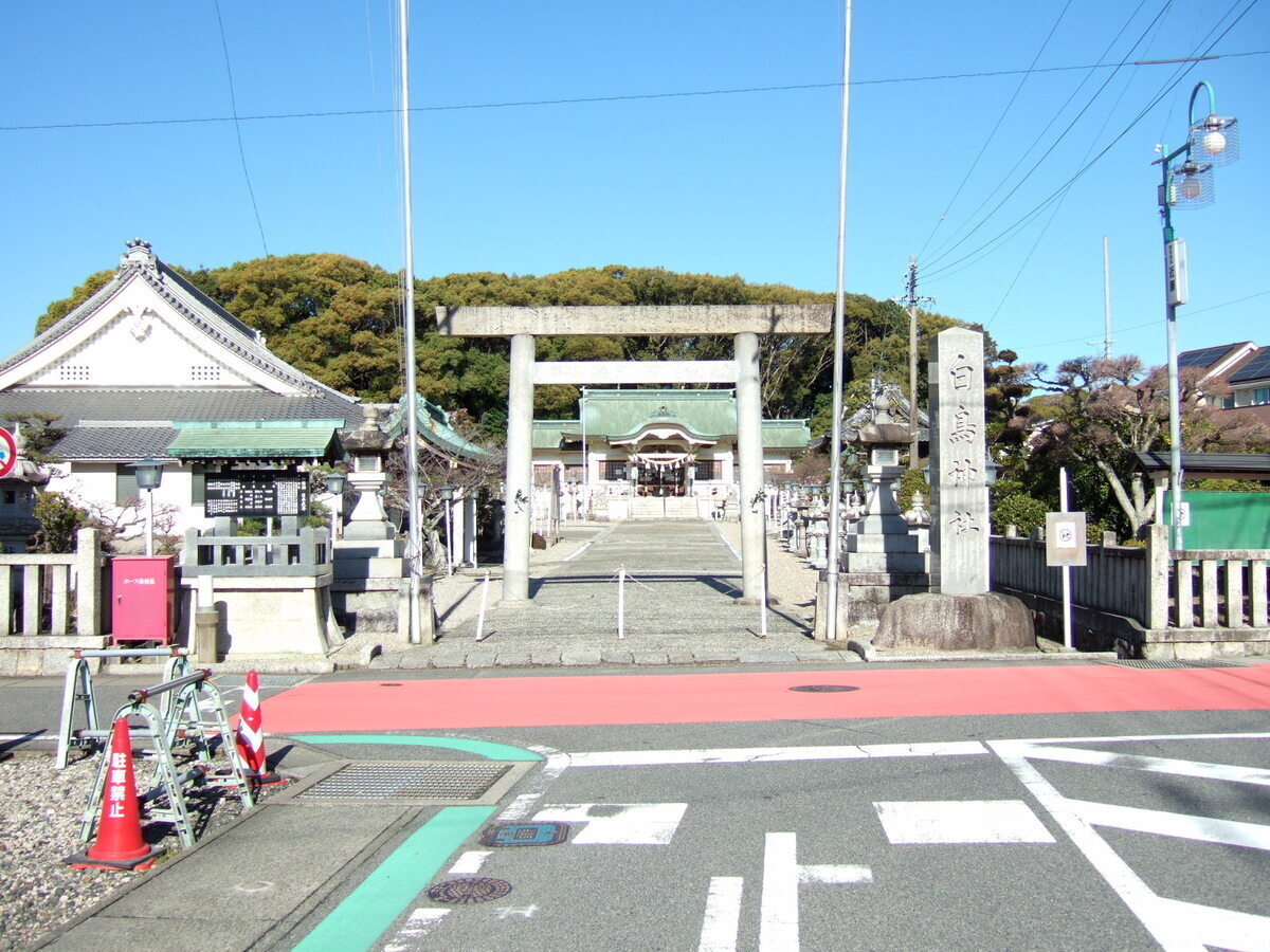 The entrance to a Shinto shrine standing at the end of a tee intersection of a suburban street. Its name (in English translation) is “White Swan Shrine.”