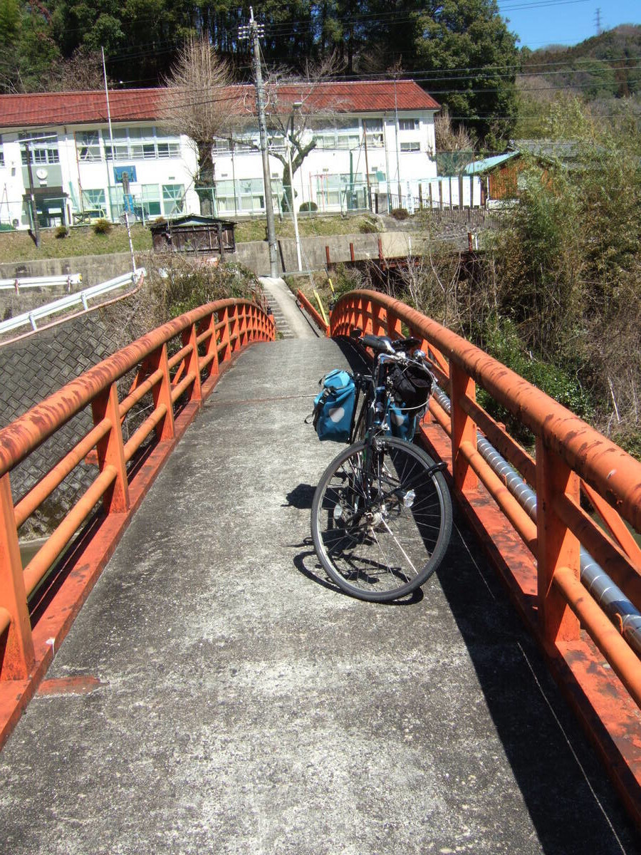 A bicycle with rear panniers parked on a steel walking bridge about 1 meter wide with faded red handrails and a concrete floor. The view looks down the bridge in the direction traveled, where a white two-story building with red roof stands across a road running perpendicular to the bridge.