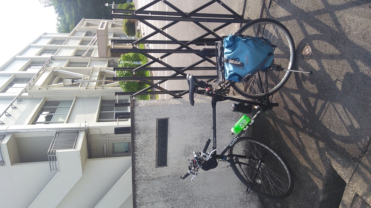 A bicycle parked in front of concrete wall and aluminum accordion gate, with a three-story concrete building in the distance.