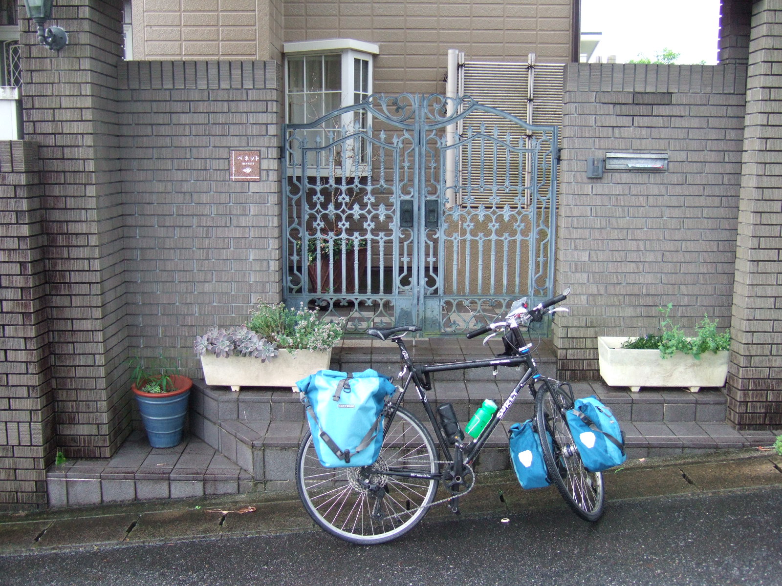 Bicycle with front and rear panniers parked at the front gate of a house. There are planters to the left and right if the gate, and in the wall to its right is a mail slot, and to its left a plaque reading “Bennett” with a small figure of two doves.