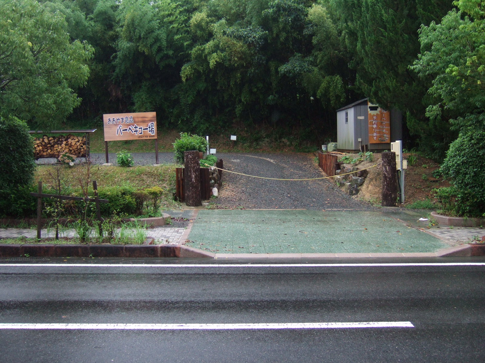 Roped-off turnout from a paved road, with a sign reading (in English translation) “Oyama Foods / Barbecue Spot”. There is a neat stack of split firewood to the left of the turn-in, with lush green foliage behind.