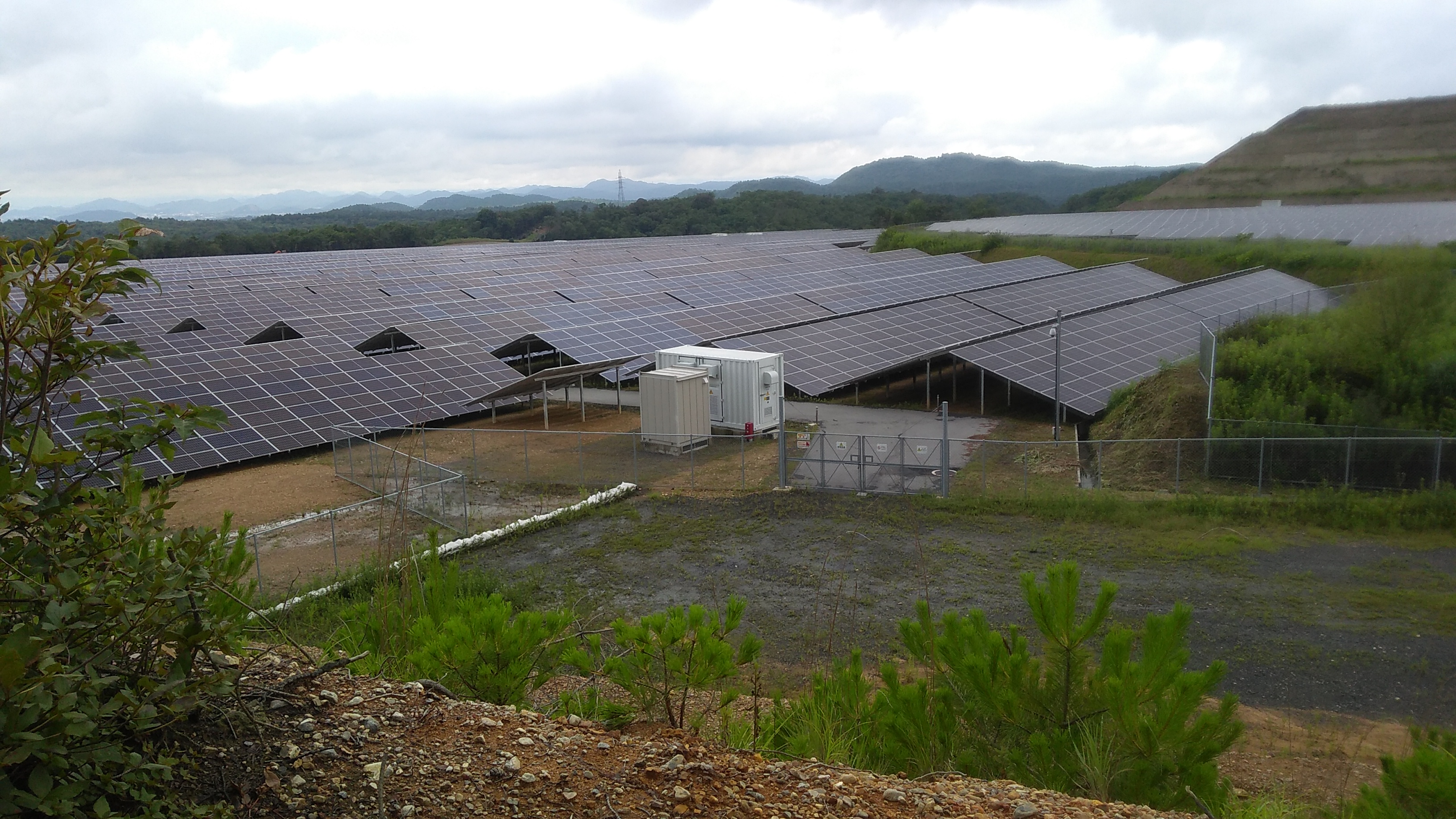 Massive expanse of solar panels in a mountain valley, with the exposed soil of a ridge terraced by earthmoving equipment visible on the horizon