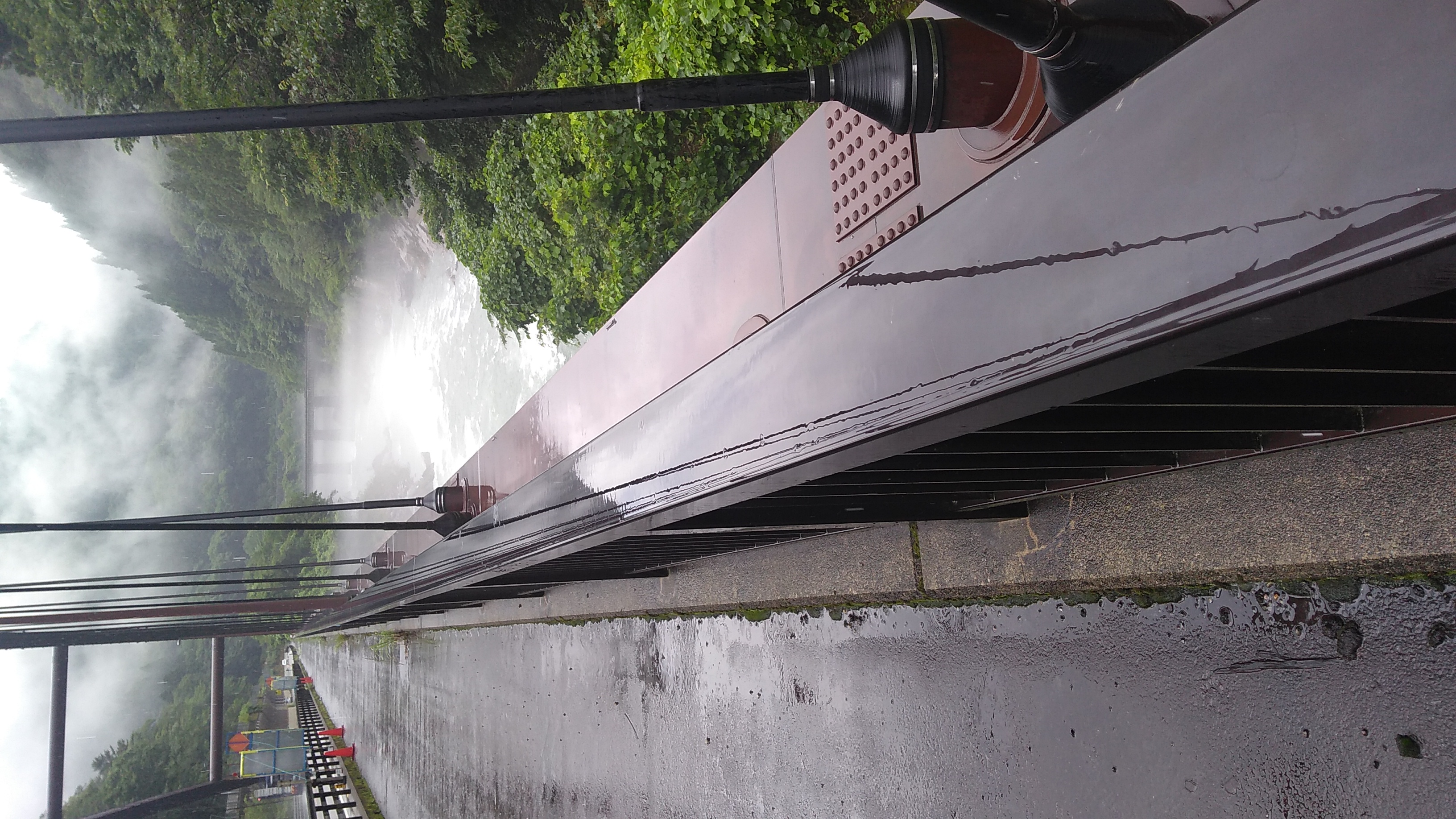 A view down the right edge of a wet steel-trussed bridge span over a river. The river bends sharply away from the camera after passing under the bridge, running in parallel with it into the distance. The far end of the river is obscured by a layer of mist lying in the gorge.