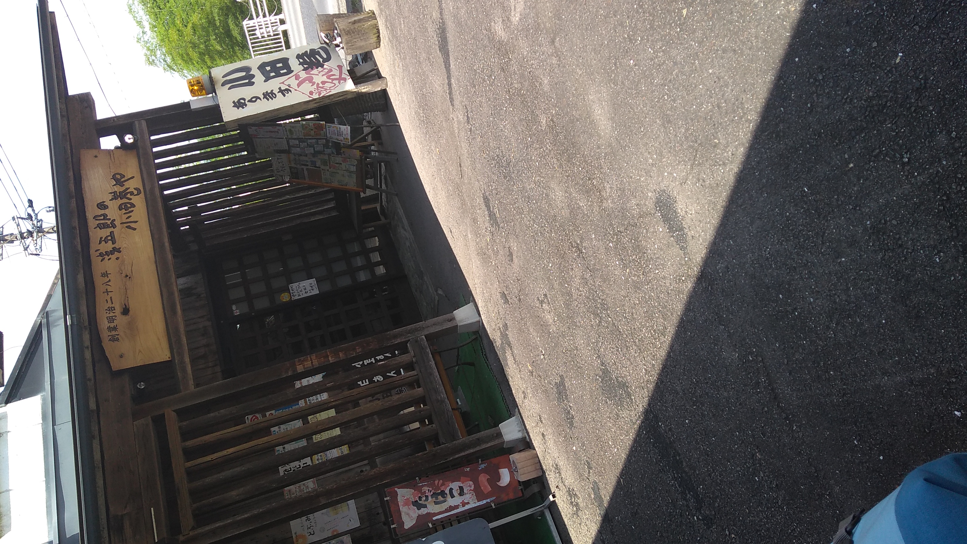 A dutch-angle view of the entrance to a shop, with a triangular patch of concreted parking lot in brilliant sunshine in the foreground. The entrance is screened lattice paneling fashioned from 2x2s on 2-inch spacing. A sign etched and stained in a rough-edged wooden slab over the entrance reads (in English translation) “Founded in Meiji 28 (1895) / Asagorō / Odamaki Vendor