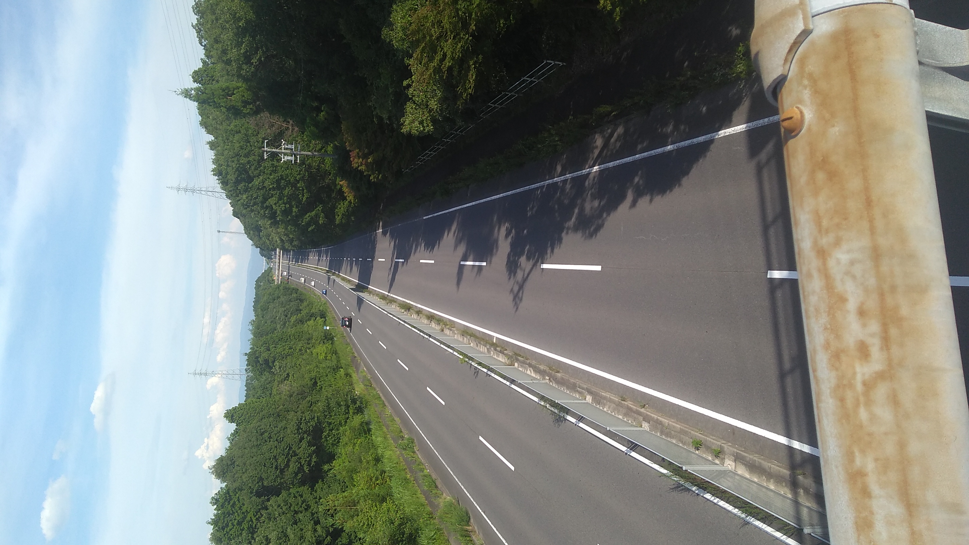 View down a four-lane expressway from the vantage point of a footbridge spanning it, with its handrail in the foreground. In the far distance, another bridge over the expressway is visible.