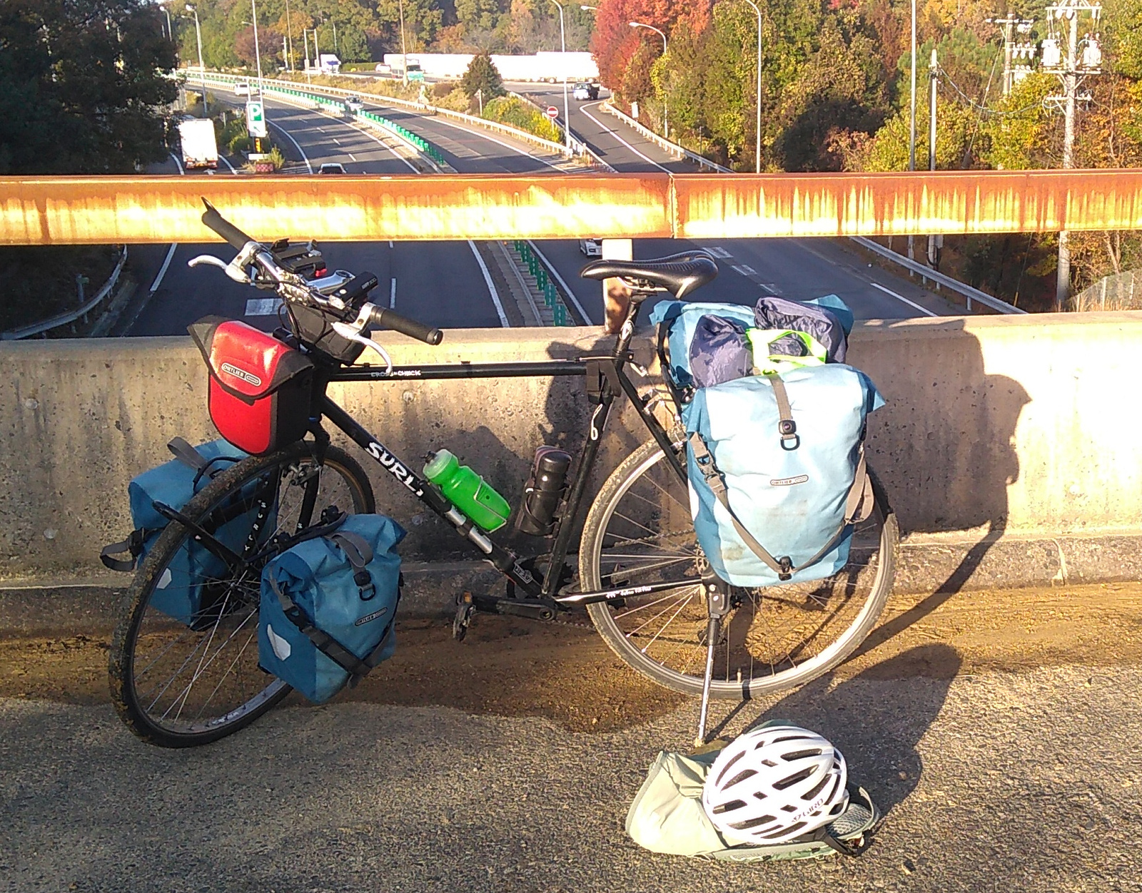 A black gravel bike parked at the rusty railing of a freeway overpass walkway, loaded up with front and rear pannier bags.