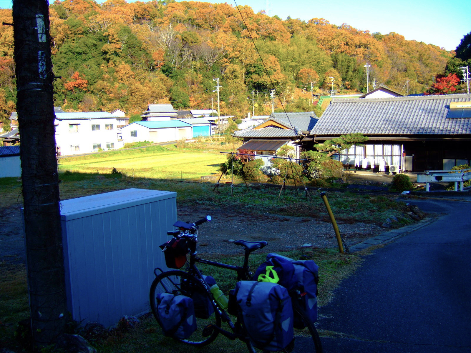 Buildings of a small mountain village, strongly lit by warm morning light cast on autumn colors of wooded hills in the background, and with deep shadow on the nearside of the scene, where a loaded touring bicycle is parked in the foreground.