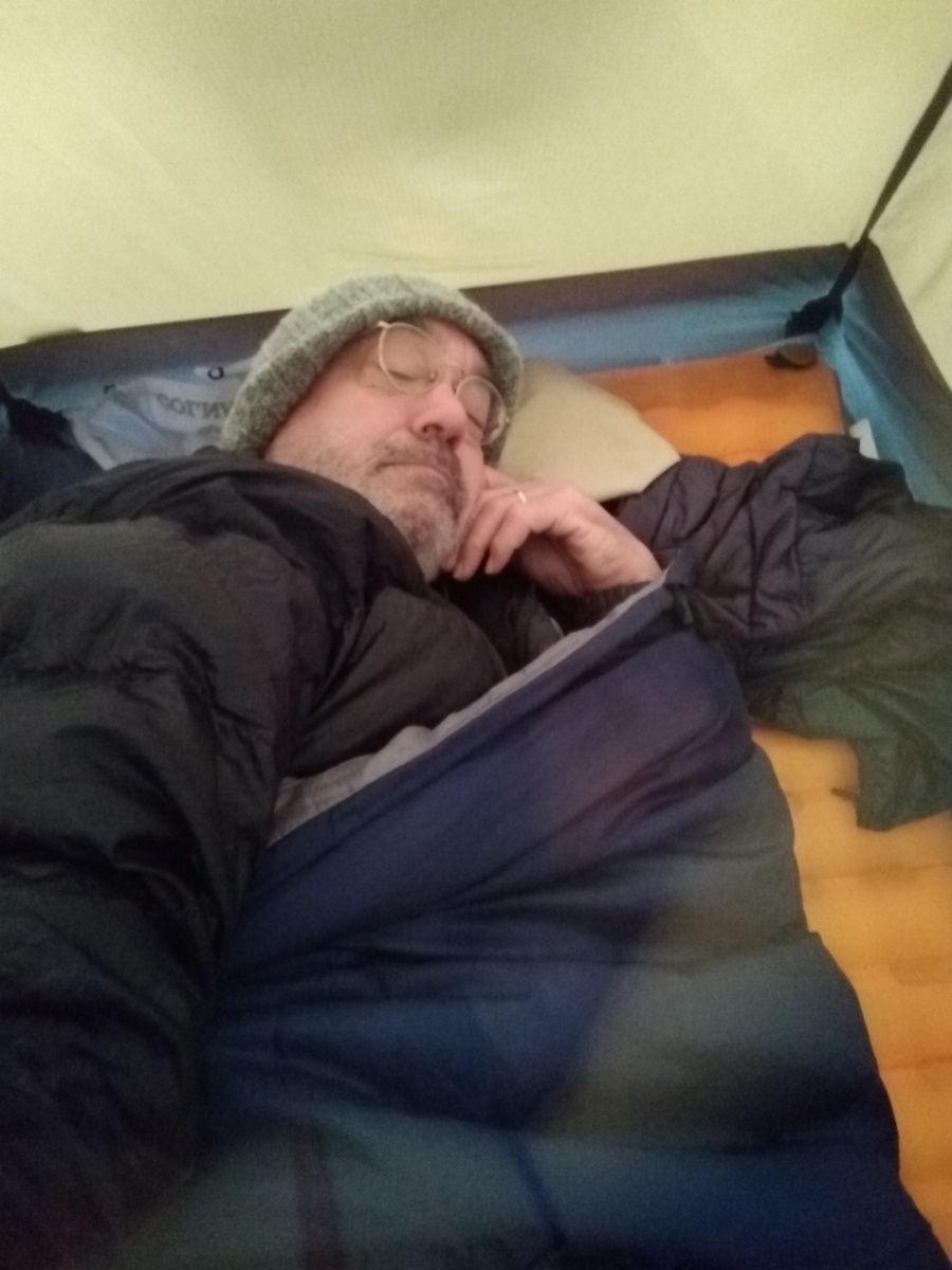 A bespectacled man with greying stubble beard, wearing a touk and squinting at the camera from inside a sleeping bag, inside a tent. His left hand bearing a wedding ring is curled against his chin.