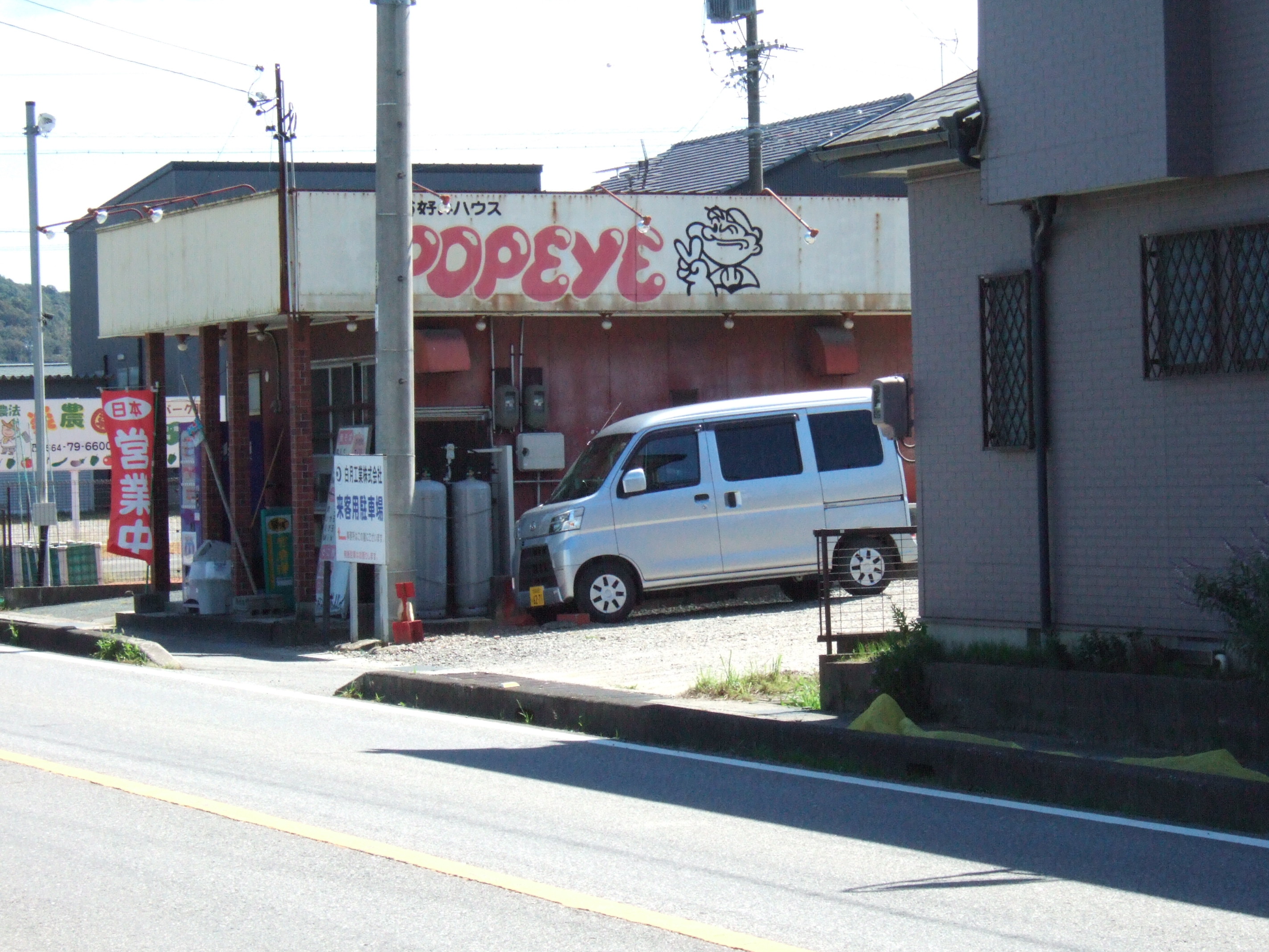 A tiny roadside restaurant nestled between concrete telephone poles with a faded sign reading “お好み焼ハウス [okonomiyaki house] POPEYE” adorned with the Popeye cartoon character flashing a peace sign.