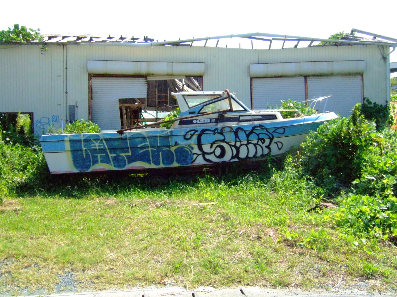 A disused motorboat covered in graffiti, sitting on a patch of grass in front of a gutted metal shed.