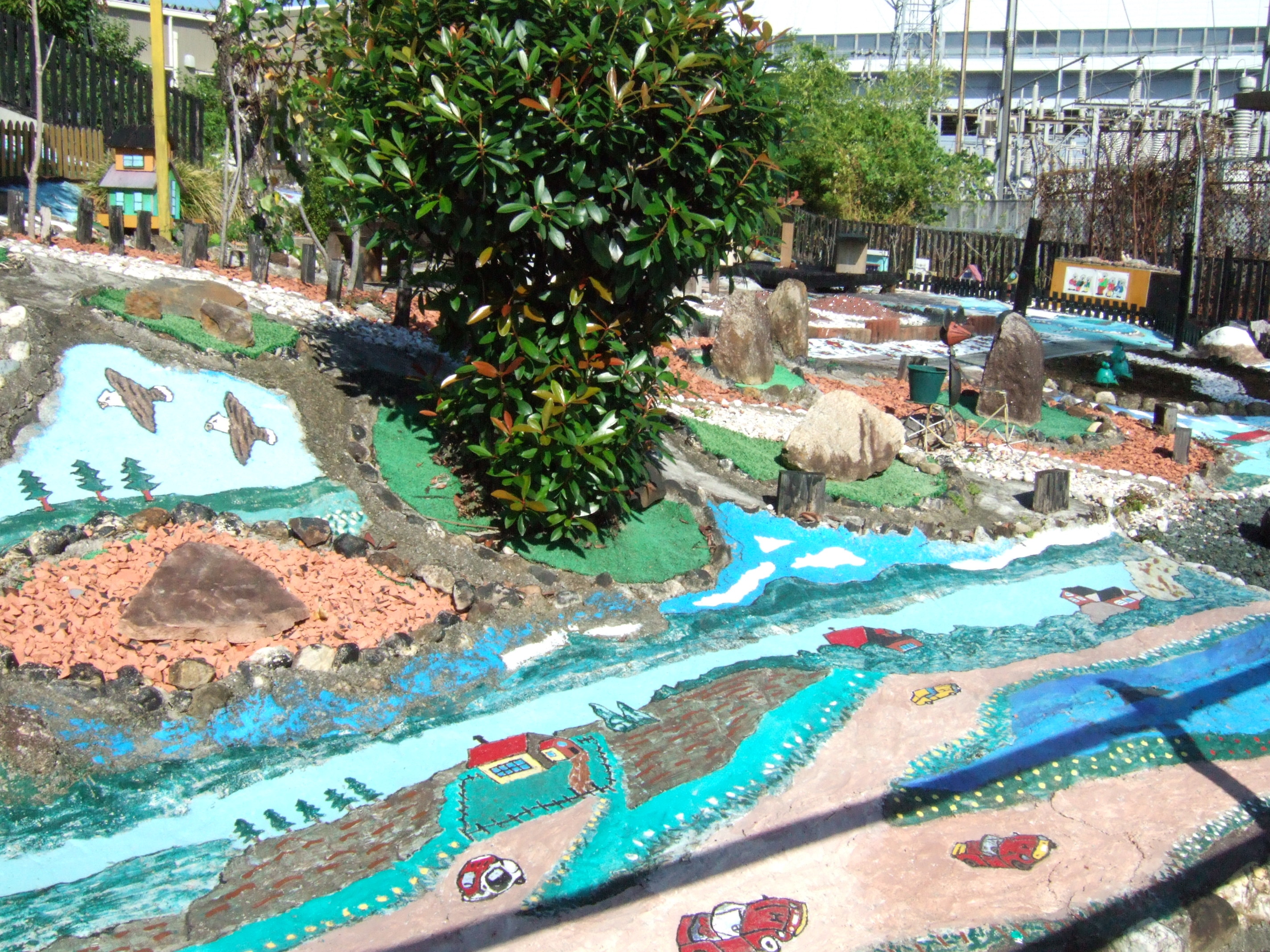 A diorama filling the entire front garden of a house. The area has been covered in cement or plaster, and decorated with some miniatures, and with streams painted in blue, and many other features painted directly onto the cement.