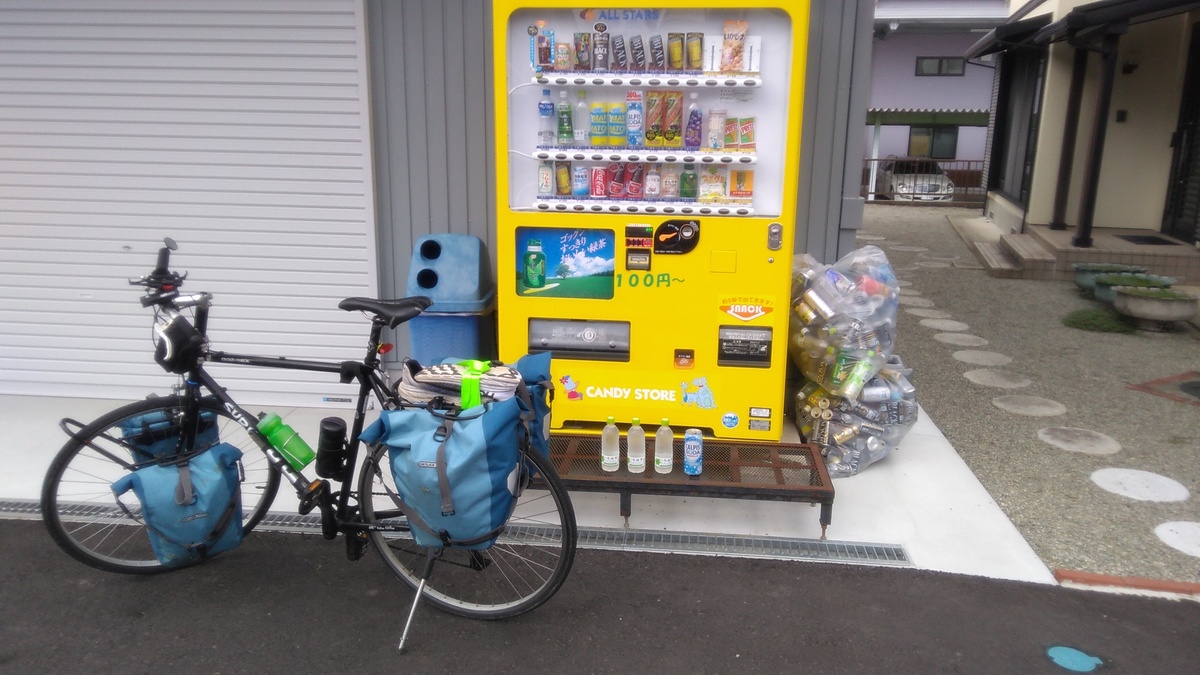 A bright yellow vending machine for drinks, with a bicycle loaded with blue panniers in front, and a row of water bottles set out beside the bike and in front of the machine.