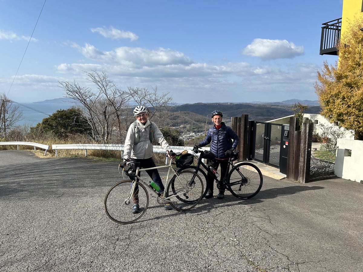 Two men in helmets, posing with their bicycles at the entry gate to a house, with a view of the horizon in the background