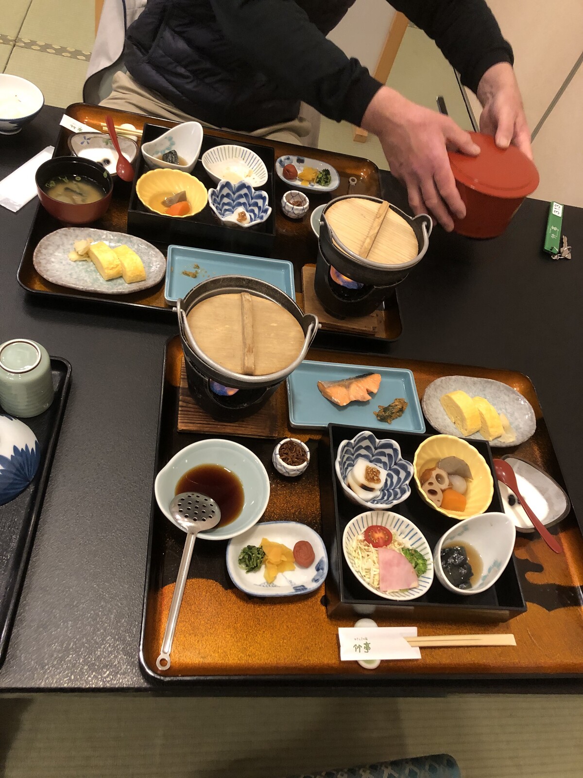 Top view of two trays, each arrayed with ten small dishes comprising a Japanese breakfast of grilled salmon, salt pickles, tofu, and other delicacies. The hands of the person sitting opposite are in frame, shifting a serving bowl of rice to the side.