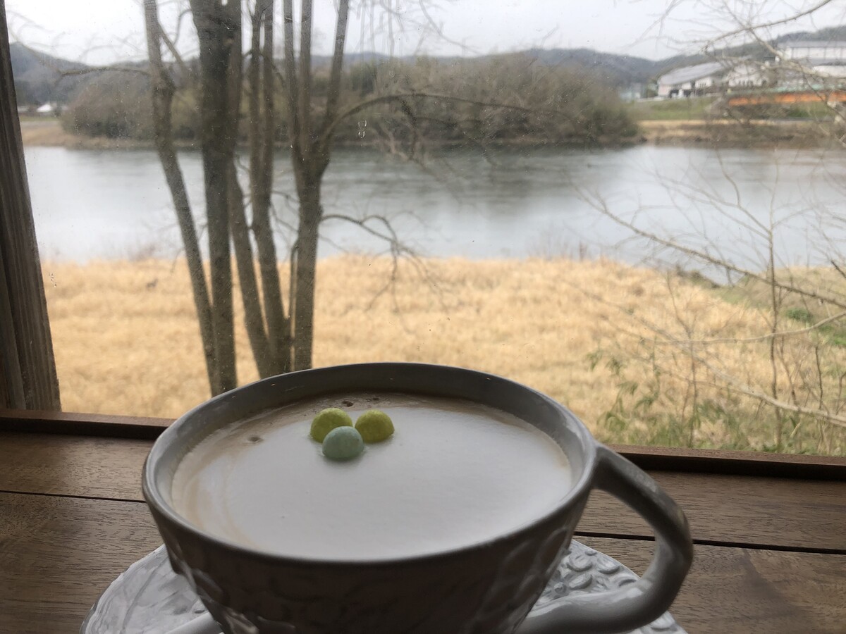 Coffee in a hand-thrown earthenware coffee cup sits on a wooden counter before a window opening onto a winter scene, and expands of dried grass on the bank of a broad river in the middle distance