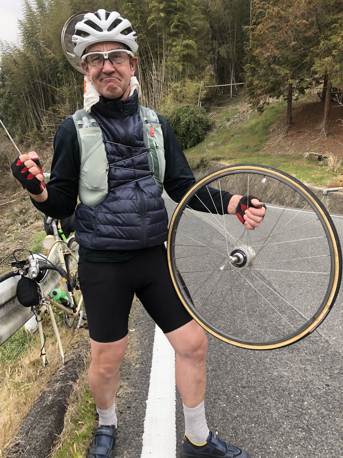 A dismounted helmeted rider, mugging for the camera while holding the uncoupled front bicycle wheel with missing spokes in one hand, and one of the spokes in the other.