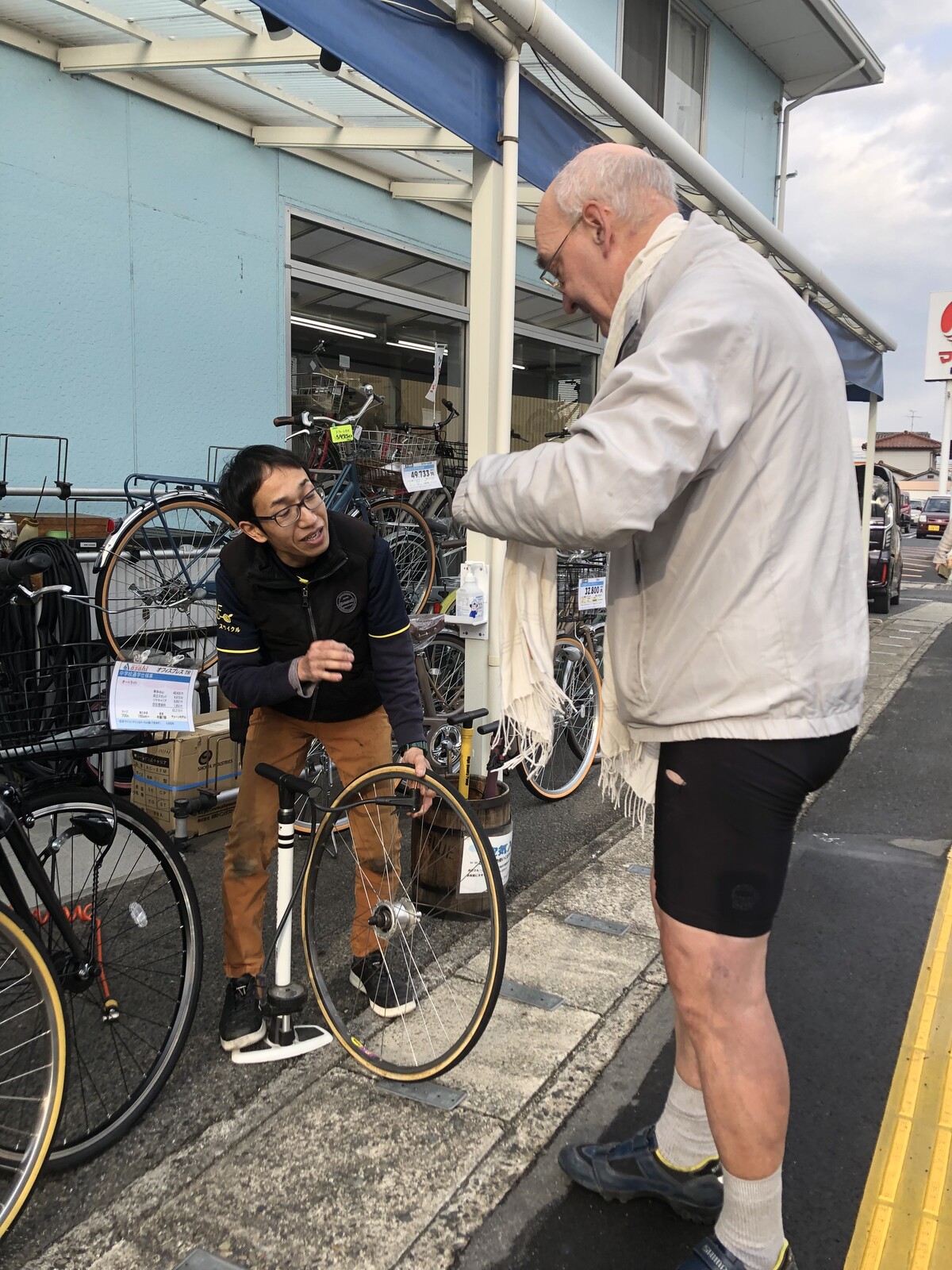 A customer (me) settling up with a mechanic in front of a bicycle shop for repair of the wheel that stands between them.