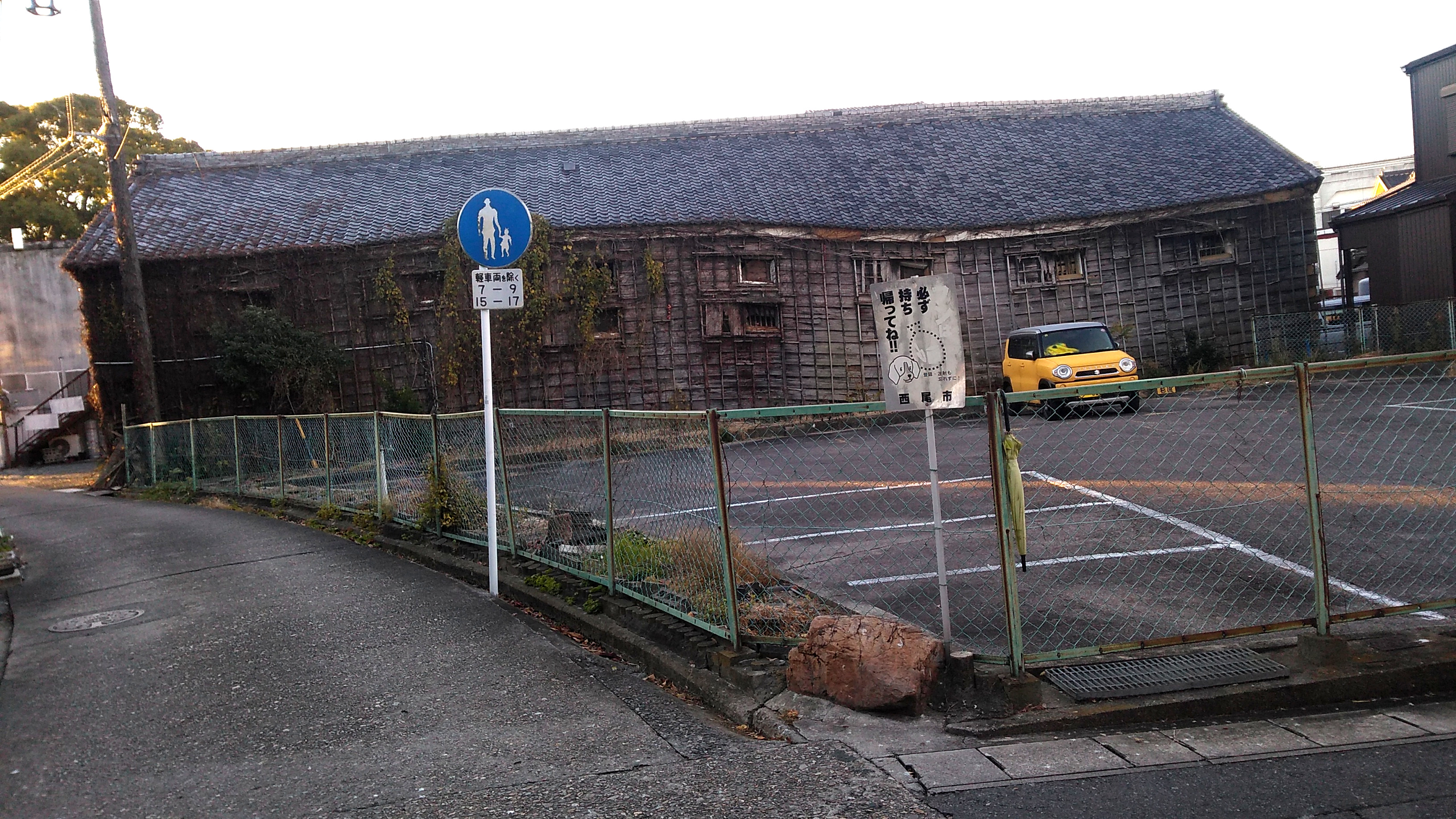 A wooden warehouse building with tile roof, viewed from the side across a parking lot behind a chain-link fence. One small yellow car is parked in front of the building. The footings of the building have subsided unevenly, twisting its lines into a wave pattern.