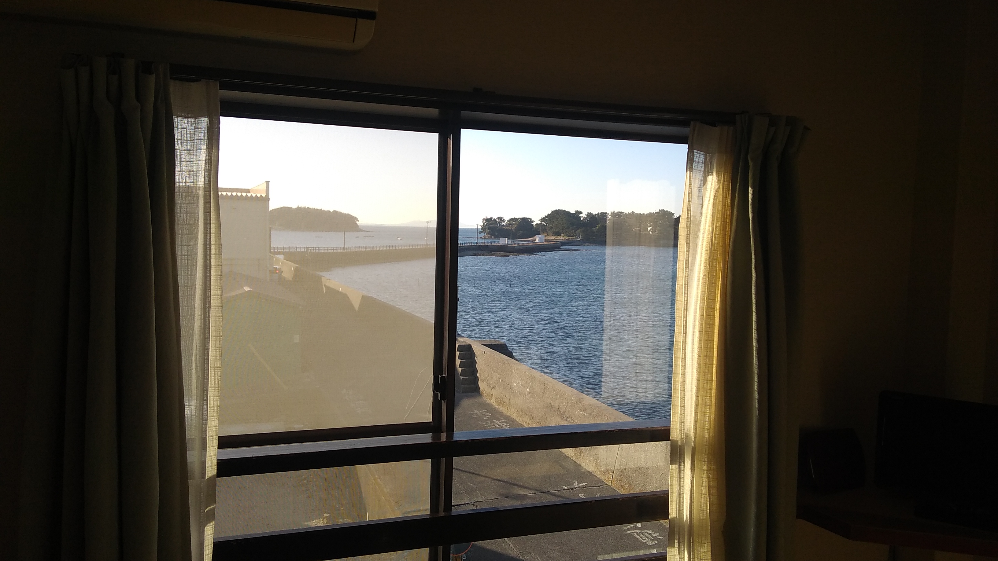 View through a second story window framed by beige curtains of a blue lagoon bounded by a sea wall, with a causeway leading to a small island visible in the right of the frame.