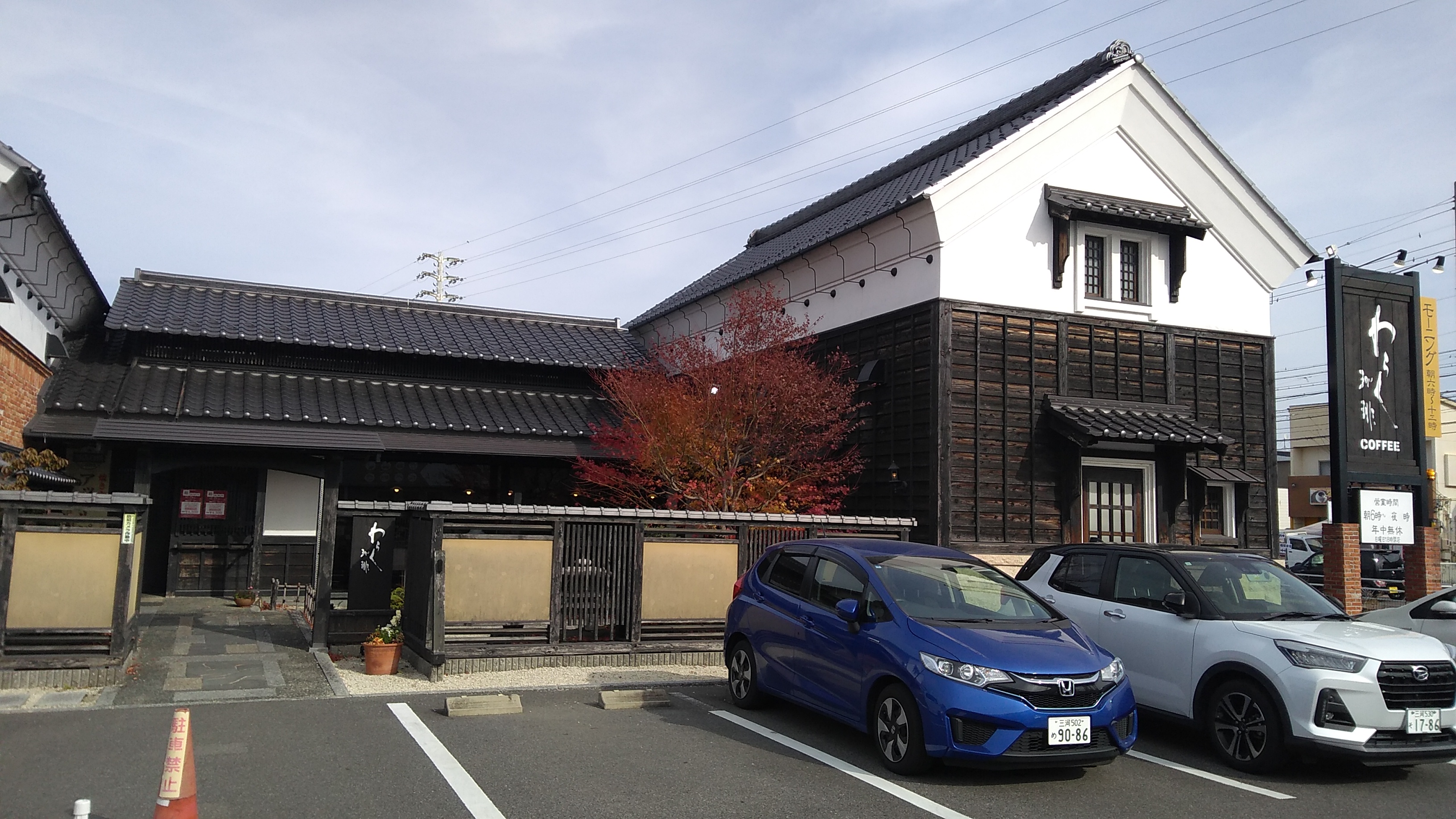 A two storey building with a peaked roof of glazed ceramic tiles and eaves finished in white plaster, with charcoal blackened cladding. A single story annex with the same finish connects this building, to the right in the frame, with a similar partner to the left.