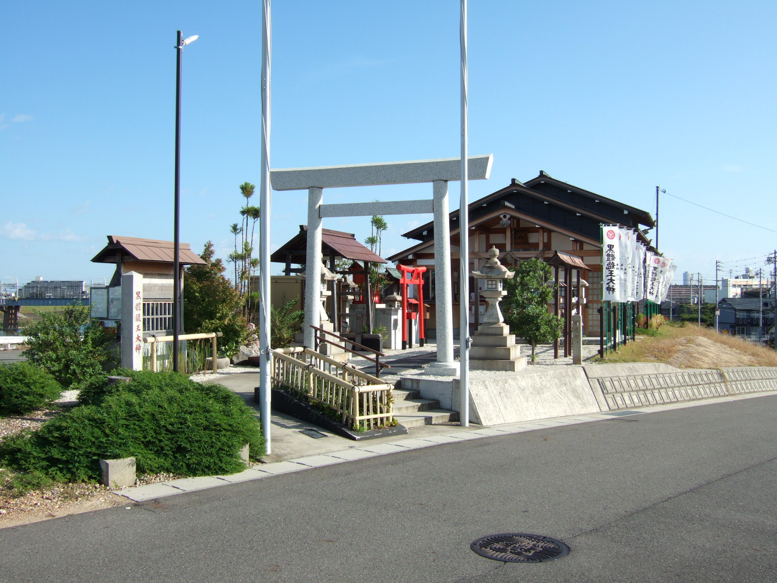 A small shinto shrine with multiple torii at the side of a road. Although not visible in the photo, the bed of a large river lies beyond the shrine.