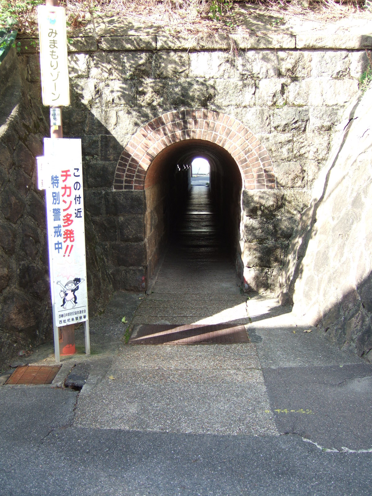 A narrow tunnel with a stonework arch through a stonework embankment. Beside the tunnel a sign reads: “Special caution: sexual assaults are common in this area!” この付近・チカン多発！！・特別警戒中 Above there is another sign reading “Protection zone” みまもりゾーン 