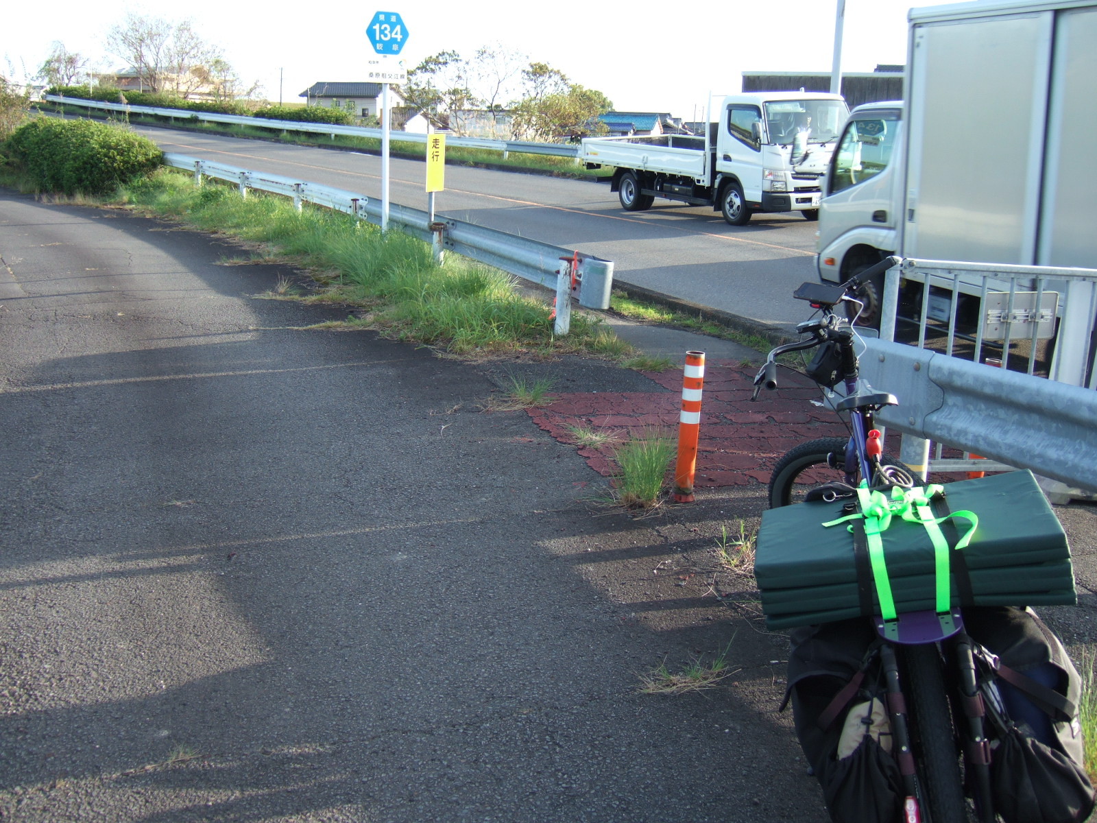 A loaded cargo bike is parked on a paved path lying alongside a busy two-lane road with trucks moving in both directions. A guard rail separates the path from the road, but scarcely noticeable is an opening to enter a narrower path on the road side of the guard rail, separated from traffic by a low concrete curb.