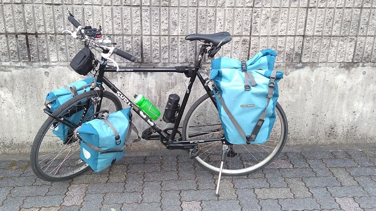 A touring bicycle with front and rear panniers parked on a brick walk in front of a tiled concrete wall.