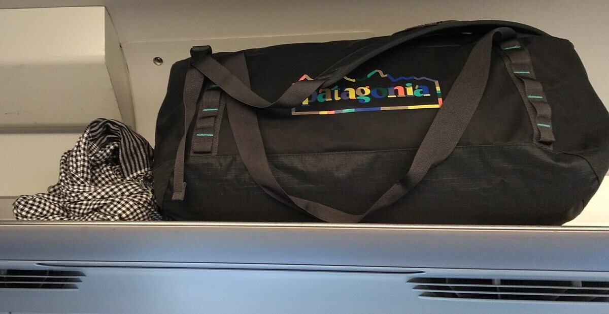 A 55-liter Patagonia duffle bag in the overhead rack of a Shinkansen carriage. A chequered cotton scarf is piled up to the left of the bag.