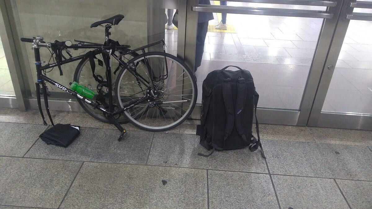 A bicycle broken down for transit with the travel cover removed, standing next to a 55-litwr duffle bag outside the Nagoya shinkansen station.